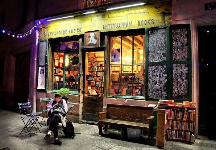 Cozy and grandiose bookstore at night with warm lighting, filled with books and a person reading outside.