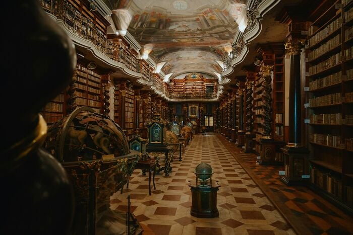 Grandiose library interior with antique globes, ornate columns, and rows of old books on wooden shelves.