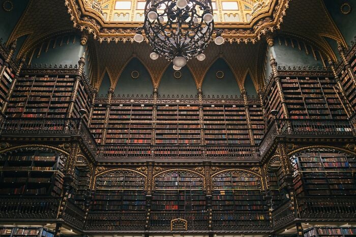 Ornate grandiose library interior with towering bookshelves and intricate gold detailing under a decorative chandelier.