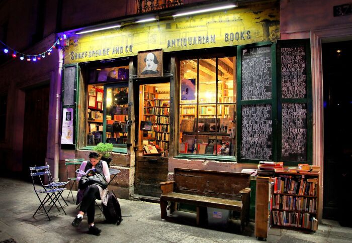 Cozy and grandiose bookstore at night with warm lighting, filled with books and a person reading outside.