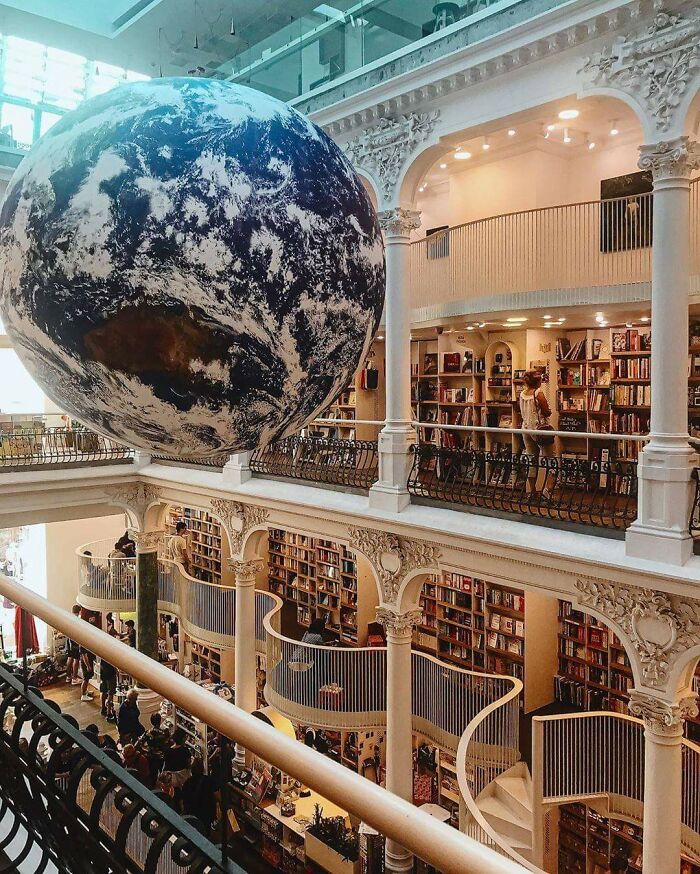 Grandiose library interior with ornate pillars, curved staircases, and a large Earth globe suspended above the bookshelves.