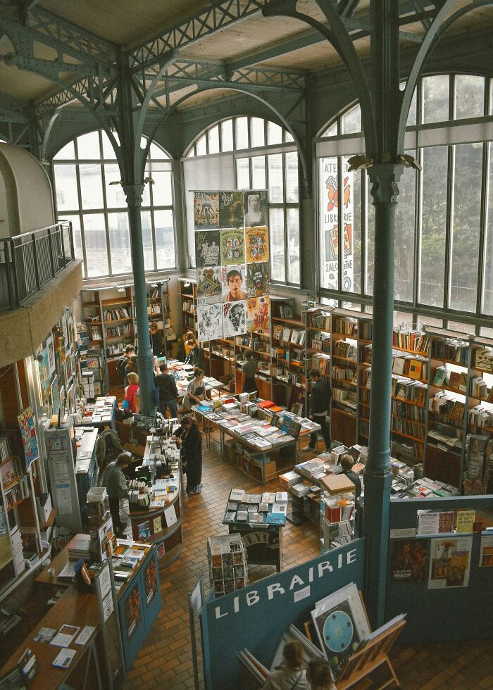Spacious, grandiose library with high arched windows, tall metal columns, and shelves filled with books and visitors browsing.