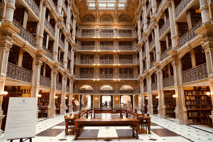 Grandiose library interior with multiple levels of bookshelves, ornate columns, and a spacious reading area with wooden tables and chairs.